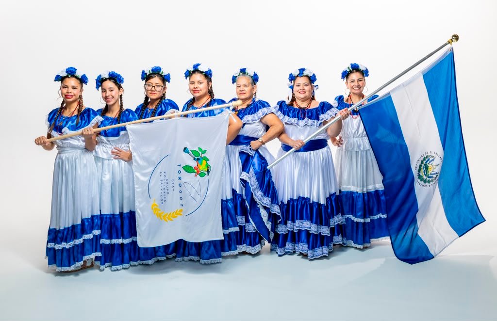 Integrantes del grupo folclórico Torogoz con trajes azul y blanco sosteniendo la bandera de El Salvador